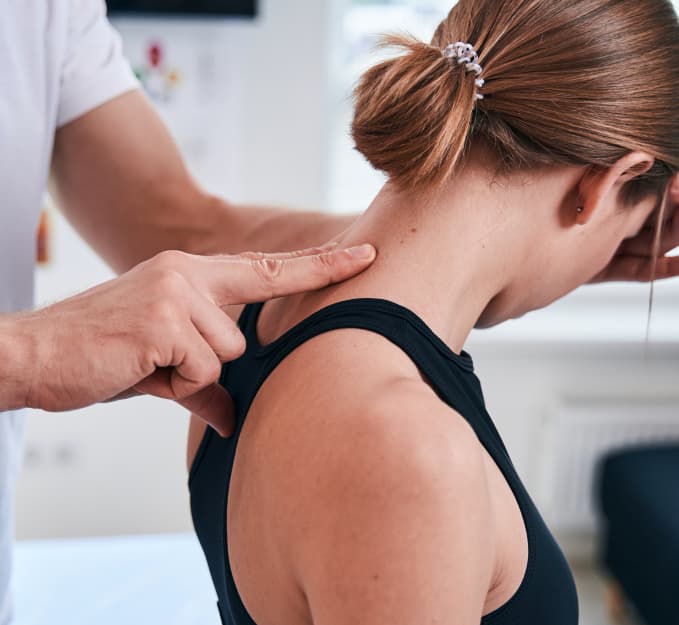 Paciente mulher em uma sala de consultório. O especialista está com dois dedos pressionando a região da nuca. A mulher usa uma regata e cabelo amarrado.